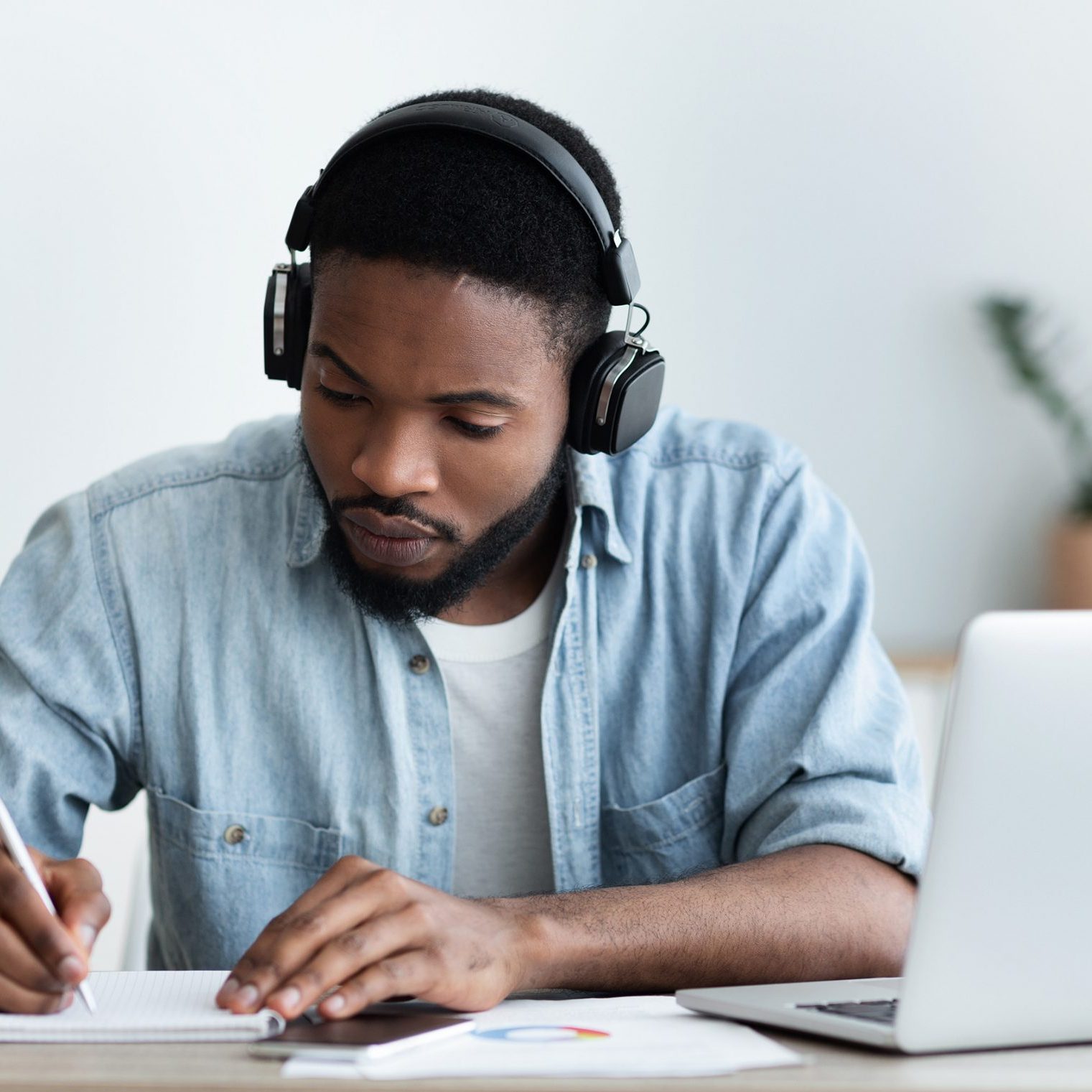 Concentrated african american guy in headphones studying foreign language online through video conference and making notes in notebook, panorama with copy space